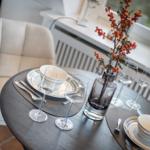 Runder Tisch mit Geschirr, Gläsern und Blumenarrangement vor Fenster.
