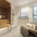 Sauna with wooden benches and glass wall. Adjacent room with chair and window.