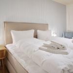 Bed with white linens and folded towels. Wooden nightstand with lamp. Striped wall.