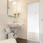 Badezimmer mit Waschbecken, Spiegel und Toilette. Tür führt in Duschbereich.