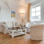 Wohnzimmer mit Sofa, Sessel und Tisch. An den Wänden hängen Bilder. Durch Fenster fällt Licht.