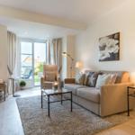 Living room with sofa, armchair, and glass table. Window to terrace. Wall art and lighting.