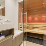 Sauna mit Holzbank und Glaswand. Neben dem Waschbecken mit Spiegel.