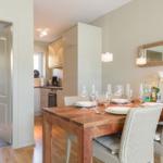 Dining area with wooden table and chairs, kitchen in background, mirror on wall.