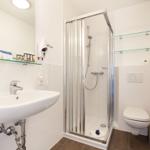 Bathroom with shower, sink, and toilet. Glass shelves and towel rack.