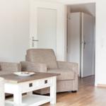 Living room with sofa, armchair, and coffee table. Kitchen and hallway are visible.