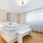 Bedroom with white bed, wooden floor, and large windows with curtains.