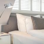 Schlafzimmer mit Bett, Nachttisch und Stehlampe unter Fenster mit Jalousie.