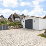 Weißes Garagenhaus mit grauen Türen und Steinmauer. Kiesweg und Hintergrundhaus sichtbar.