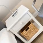 White nightstand with drawer and basket. Lamp and bed are visible.