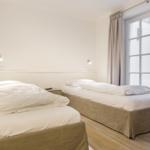 Zwei Betten mit weißen Bettwäschen und Holzboden. Fenster mit Vorhängen.