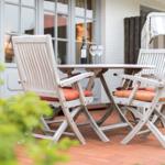 Terrasse mit Tisch, zwei Stühlen und Weinflasche. Hintergrund: Haus mit Fenstern und Sonnenschutz.