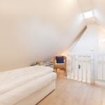 Schlafzimmer mit Dachschrägen, Bett, Stuhl und Sicht auf Treppenhaus.