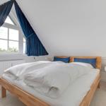 Schlafzimmer mit Holzbett, blauen Vorhängen und Fenster. Neben dem Bett steht ein weißer Nachttisch.