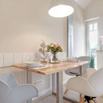 Dining area with wooden table and white chairs. Kitchen with white cabinetry and window.