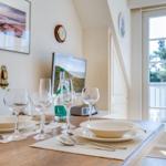 Dining table set with dishes, glasses, and bottle. Windows open to balcony with green view.