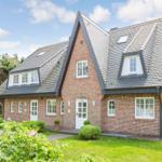 Red brick house with dormer windows and white entrance. Green lawn and trees surround the house.