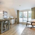 Dining area with table and chairs, living area with armchair and sofa, wooden floor, large windows.