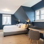 Schlafzimmer mit Bett, Schreibtisch und Fenstern.
