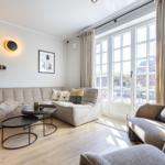 Living room with sofa, armchairs, and tables. Large windows and wooden floor.