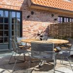 Terrace with round table and chairs in front of brick house with windows and roof.