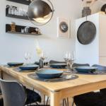 Dining table with six chairs and tableware. Background: kitchen cabinets and wall shelves.