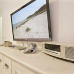 TV auf Sideboard zeigt Strandbild, daneben steht ein weißer Lautsprecher.
