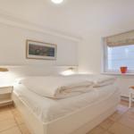 Bedroom with white bed, nightstand, and chair beside window.