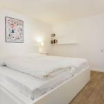 Bedroom with white bed, wooden floor, and wall art. Door leads to staircase.