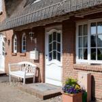 Red brick house with thatched roof, white doors and windows, flower pots and bench at entrance.
