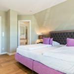Schlafzimmer mit großem Bett, lila Kissen und Holzboden. Blick in Bad und Wohnbereich.