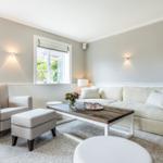 Living room with sofa, armchair, and wooden table. Windows and wall lights.