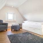Schlafzimmer mit Dachschrägen, weißem Bett und grauer Sitzgelegenheit.