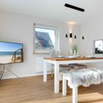 Essbereich mit Tisch, Stühlen und Fernseher. Fenster zeigt Haus und Strand.
