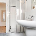 Bathroom with shower, sink, and mirror. Corridor leads to a room with mirror.