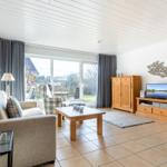Living room with sofa, coffee table, and TV. Large windows with garden view.