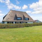 Haus mit Strohdach, umgeben von Gras und Hecken unter blauem Himmel.
