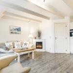 Wohnbereich mit Sofa, Kamin und Küchenzeile. Holzboden und weiße Einrichtung.