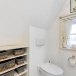 Badezimmer mit Toilette, Regal und Fenster mit Vorhang.