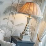 Table lamp on nightstand beside bed, wall with palm pattern, white curtains.