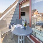Terrasse mit Tisch, zwei Stühlen und Flasche. Blick auf Wasser und Gebäude.