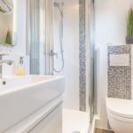Badezimmer mit Dusche, Waschbecken und Toilette. Weiße Fliesen und Mosaikverkleidung.