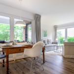 Dining area with wooden table and white chairs. Living area with sofa and armchairs. Large windows with garden view.