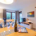 Dining area with wooden table, wine bottle, and book. Background: living room with TV and glass cabinet.