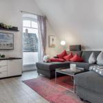 Wohnzimmer mit Sofa, Fernseher und Fenster. Auf dem Sofa liegen Kissen und eine Decke.