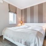 Schlafzimmer mit Bett, Nachttisch und Lampe. Wand mit Streifenmuster. Fenster mit Rollladen.