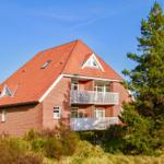 Ein zweistöckiges Haus mit rotem Dach und weißen Balkonen. Umgeben von Gras und einem großen Baum.