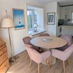 Round table with four pink chairs, beside wooden cabinet and lamps. Kitchen in background.