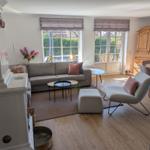Living room with sofa, armchair, and dining table. Wooden floor and large windows.