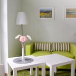 Ein Wohnzimmer mit grünem Sofa, weißen Tischen und Fenster. Wandbilder und Stehlampe.